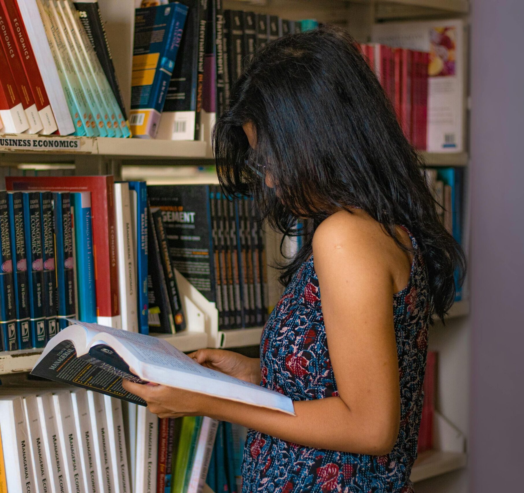 Young woman reading a book in a library in Delhi, India, surrounded by shelves of business economics books.